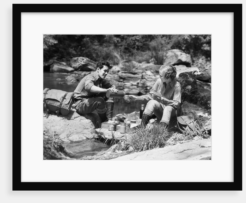 Couple having picnic sitting on rocks by stream man pouring drink for woman from thermos by Anonymous