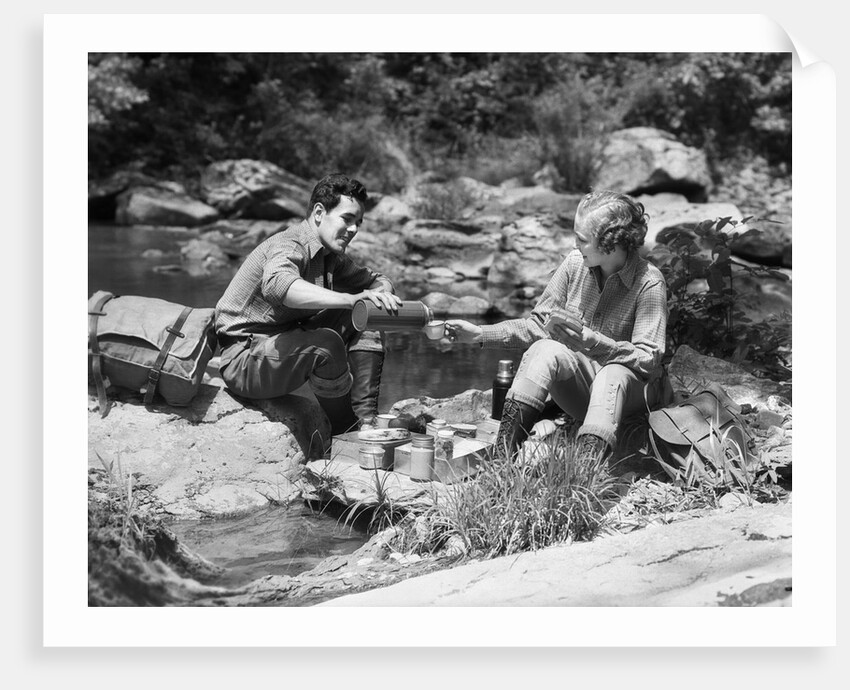 Couple having picnic sitting on rocks by stream man pouring drink for woman from thermos by Anonymous