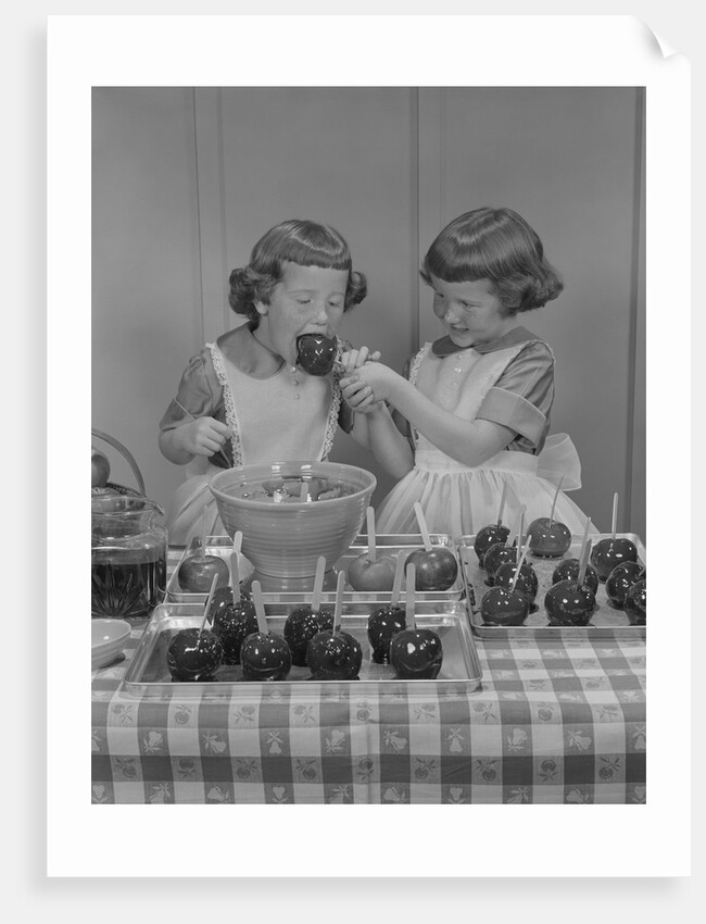 Two twin girls eating making candy apples in kitchen by Anonymous