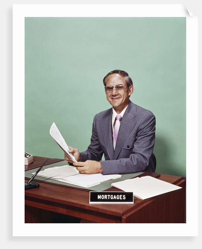 Smiling man at desk holding papers with mortgage officer sign looking at camera by Anonymous