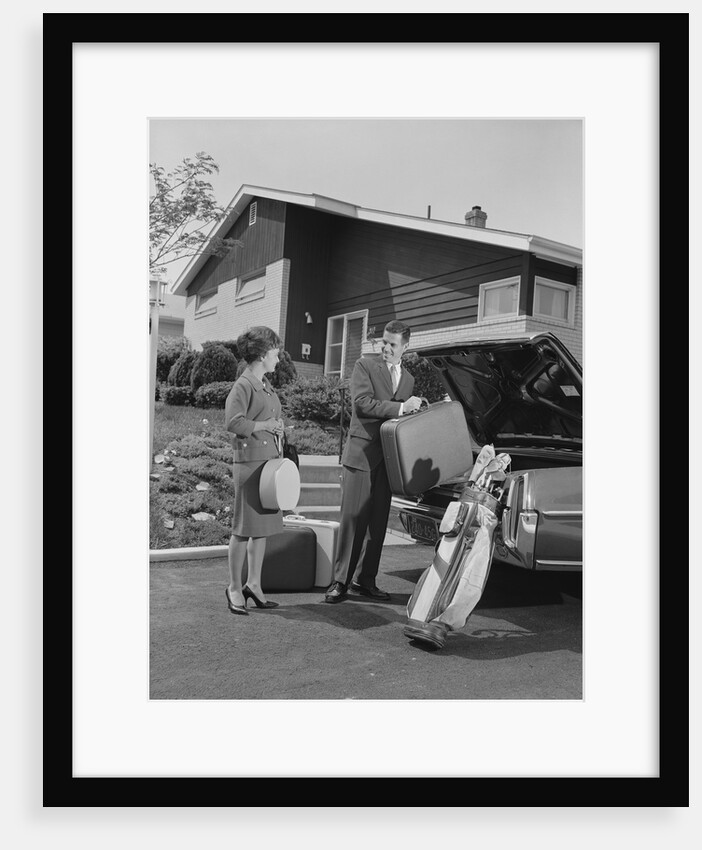 Couple man woman packing luggage into car trunk by suburban house by Anonymous