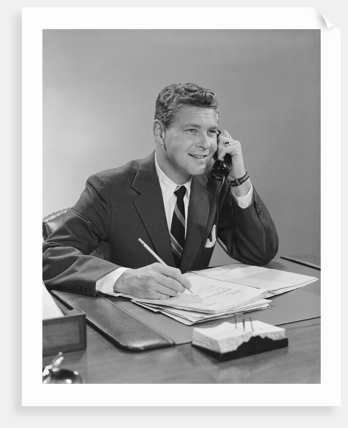 Businessman seated at desk talking on telephone smiling writing with pencil by Anonymous