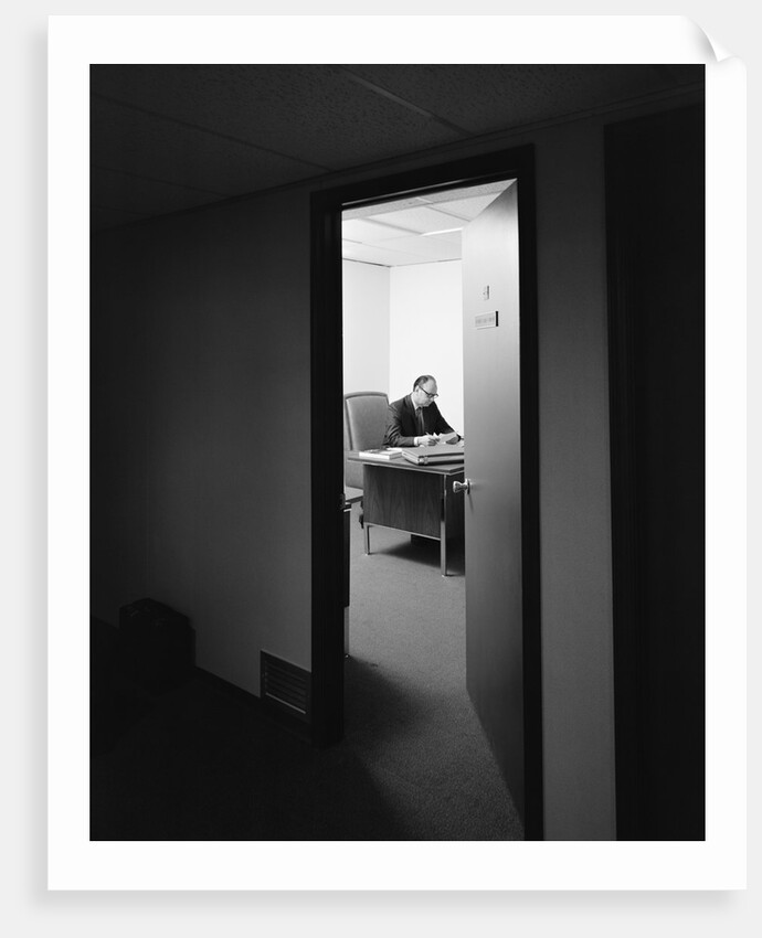 Executive businessman at desk seen working late through door ajar by Anonymous