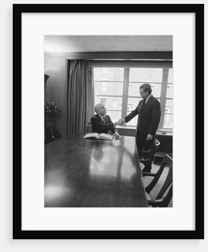 Two men one seated shaking hands long wooden conference table by Anonymous