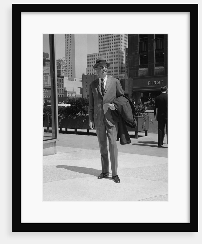 Businessman wearing suit hat carrying top coat standing on city street by Anonymous