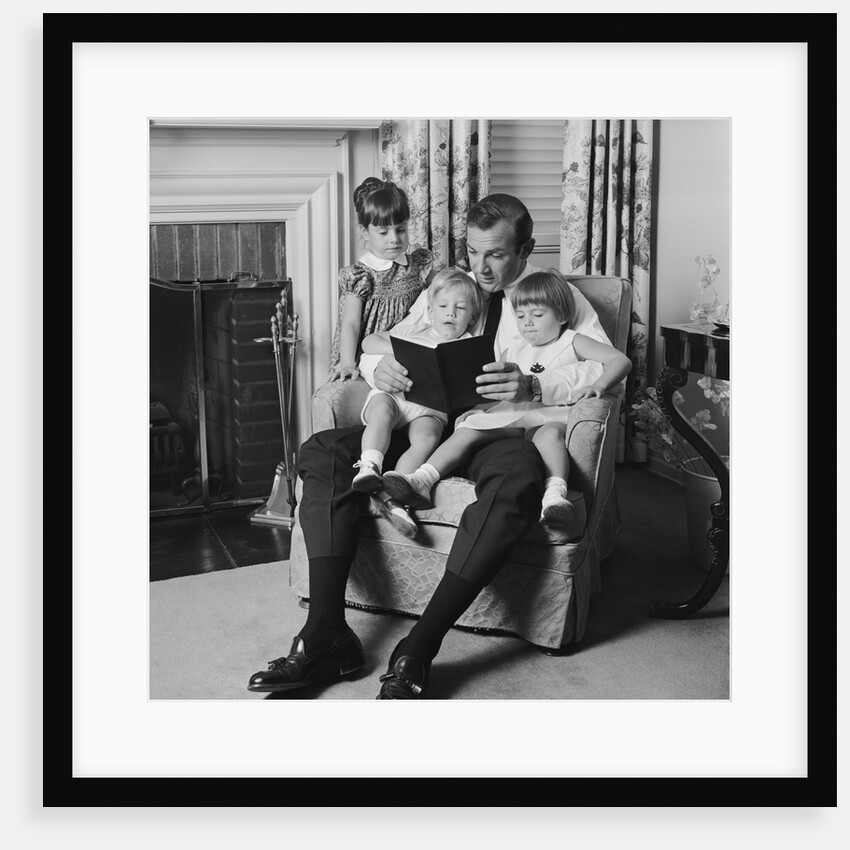 Man father sitting in chair with three children reading book by Anonymous