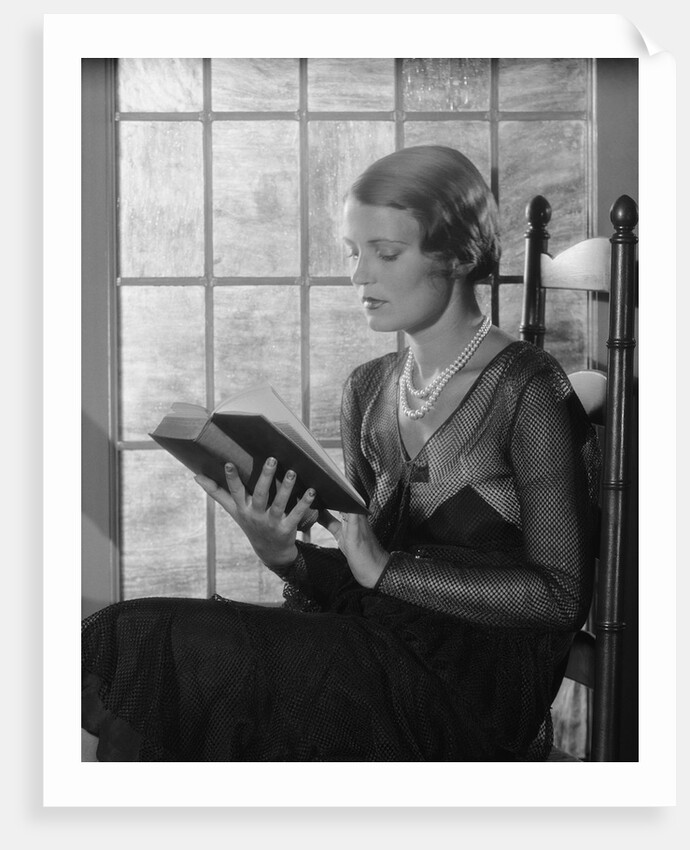Elegant woman wearing pearls sitting by window reading book by Anonymous