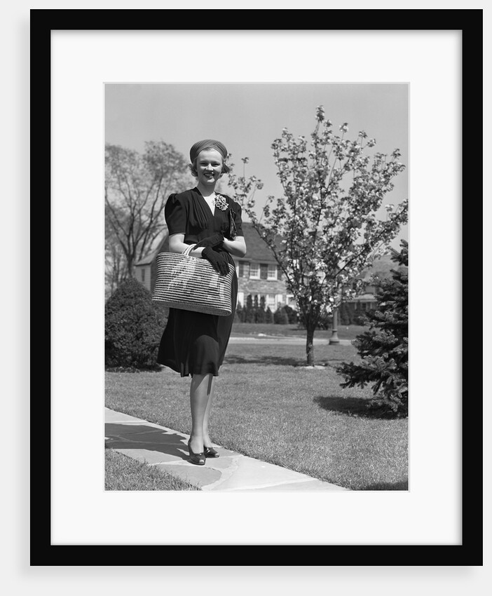 Stylishly dressed woman walking down suburban street carrying straw bag by Anonymous