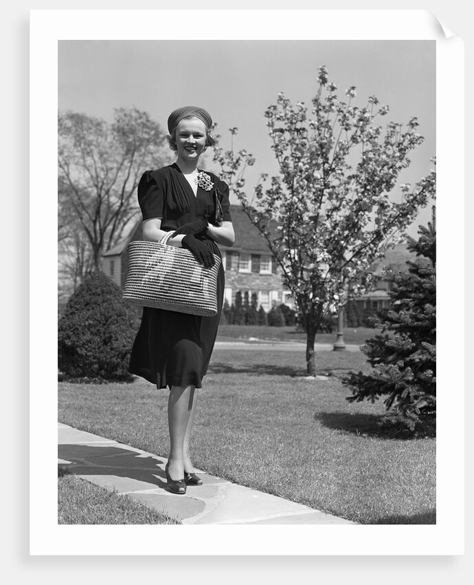 Stylishly dressed woman walking down suburban street carrying straw bag by Anonymous