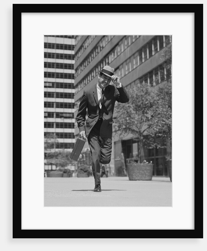 Businessman carrying briefcase holding his hat on running down urban sidewalk by Anonymous