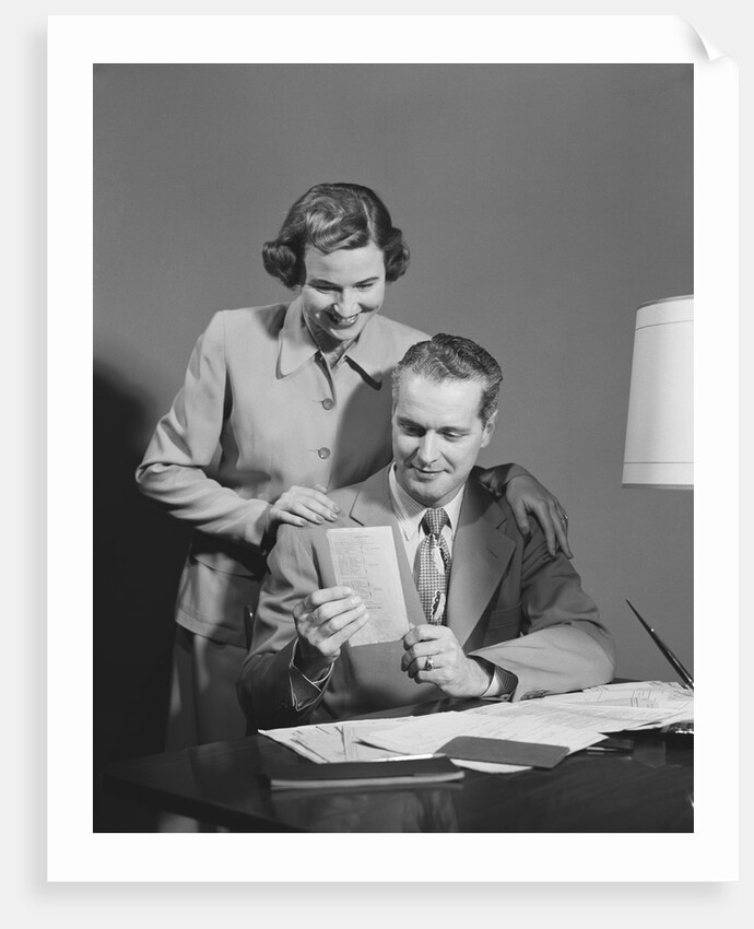 Couple man seated at desk holding paper woman standing looking over his shoulder smiling happy by Anonymous
