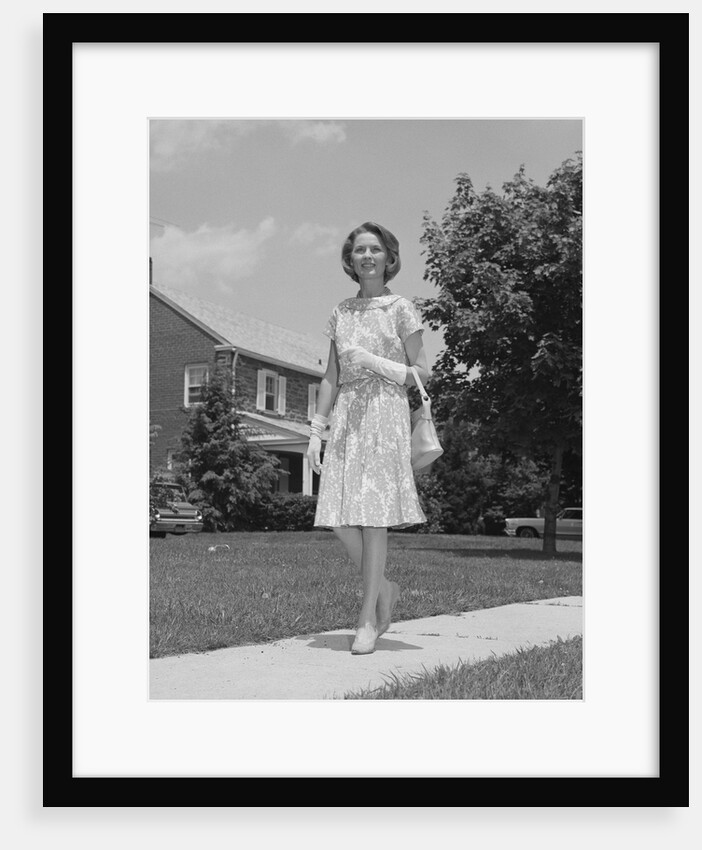 Woman wearing print dress holding handbag walking down suburban street by Anonymous