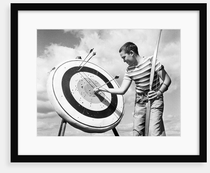 1960s boy jeans striped t-shirt holding bow & pulling arrow out of target bull's-eye by Anonymous