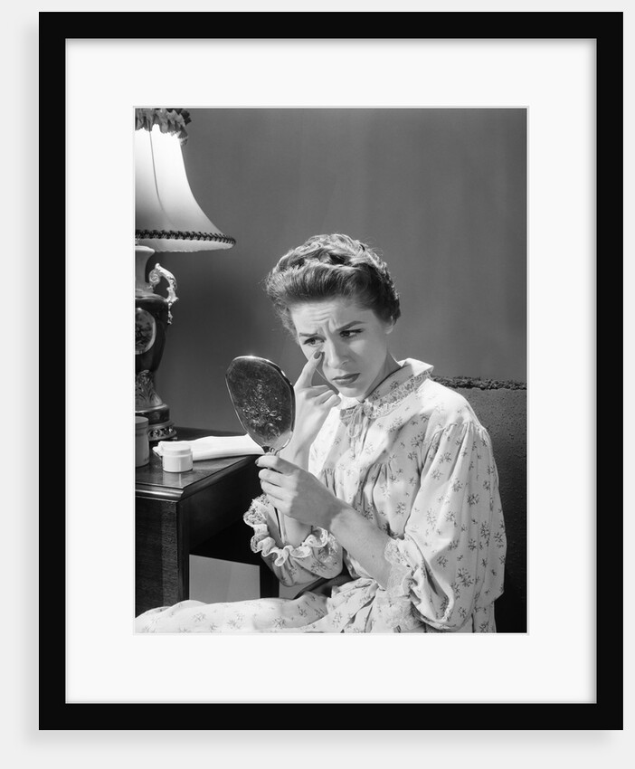 1950s 1960s woman in nightgown holding mirror look at eye wrinkles aging beauty cosmetic serious concerned expression by Anonymous