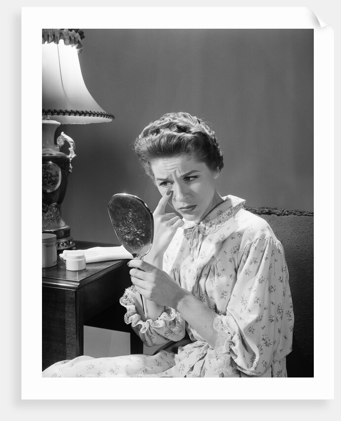 1950s 1960s woman in nightgown holding mirror look at eye wrinkles aging beauty cosmetic serious concerned expression by Anonymous