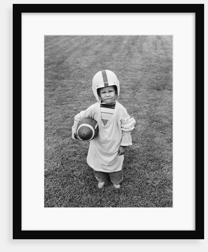1950s boy standing in grass wearing oversized shirt & helmet holding football by Anonymous