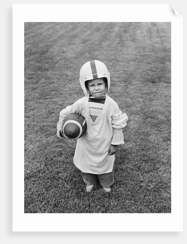 1950s boy standing in grass wearing oversized shirt & helmet holding football by Anonymous