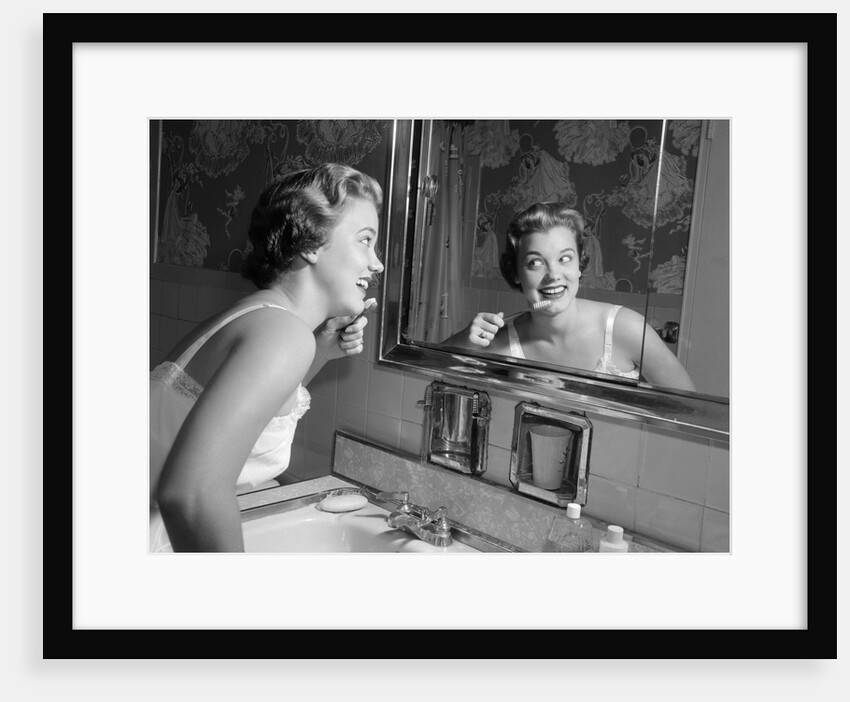 1950s smiling woman looking into bathroom mirror brushing teeth by Anonymous