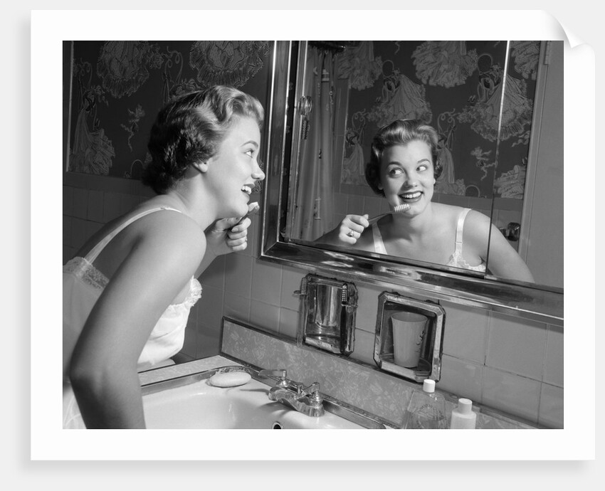1950s smiling woman looking into bathroom mirror brushing teeth by Anonymous