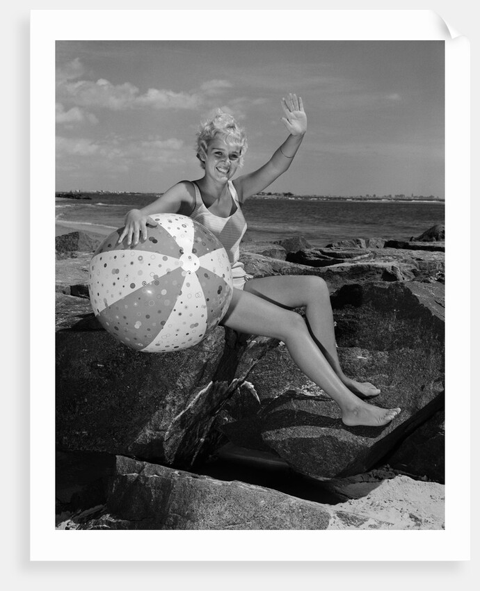 1960s smiling blond teenage girl sitting on rocky shore holding a beach ball waving looking at camera by Anonymous