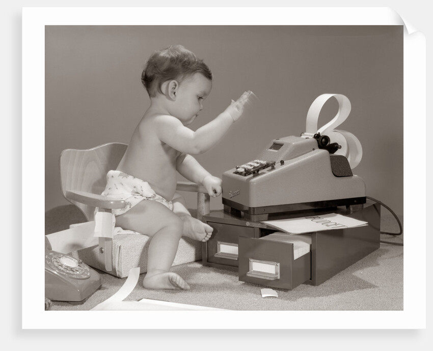 1960s baby seated in small chair hitting keys on office adding machine on top of small file drawers by Anonymous