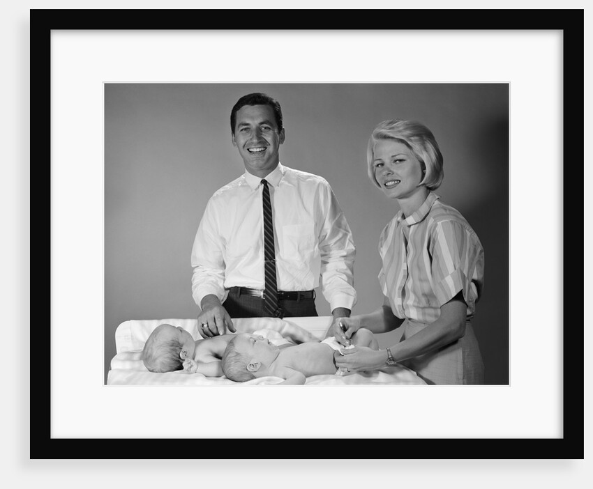 1960s smiling mother and father with twin babies on diaper changing table looking at camera by Anonymous