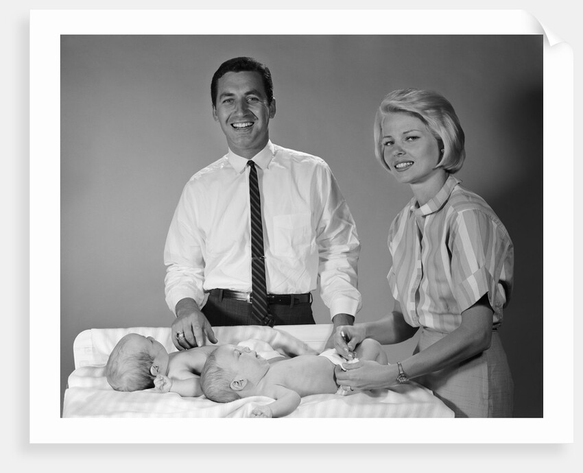 1960s smiling mother and father with twin babies on diaper changing table looking at camera by Anonymous