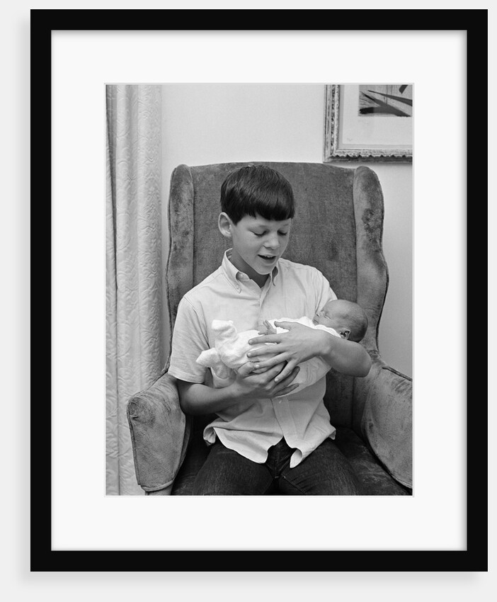1960s pre-teen boy sitting in armchair holding infant newborn baby sibling by Anonymous
