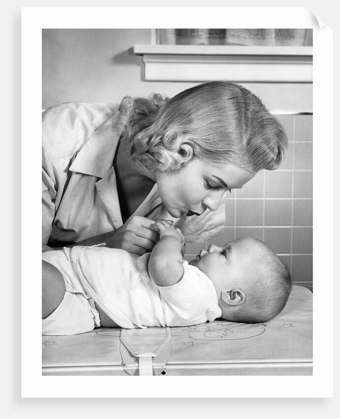 1950s mother woman bending over baby lying on layette holding hands talking by Anonymous