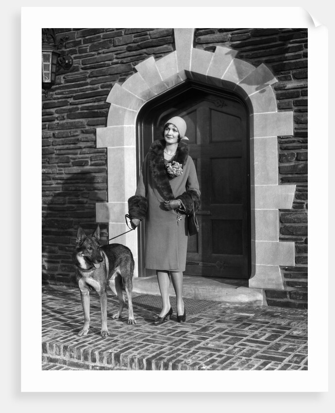 1920s fashionable smiling woman wearing cloche hat coat with fur trim and german shepherd dog on leash standing in arched doorway by Anonymous
