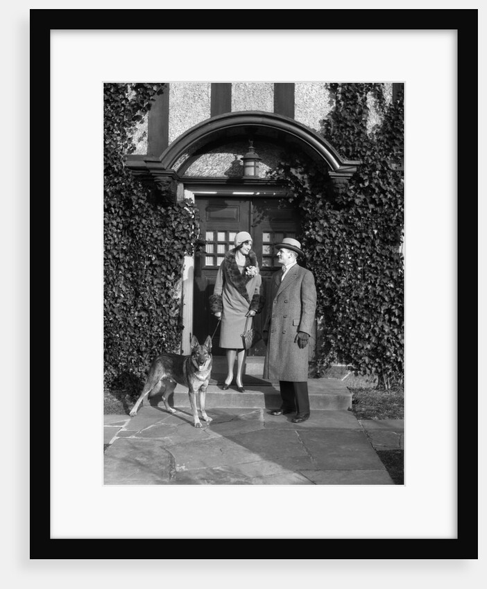 1920s couple wearing coat hat gloves on steps ivy covered building with german shepherd dog on a leash by Anonymous