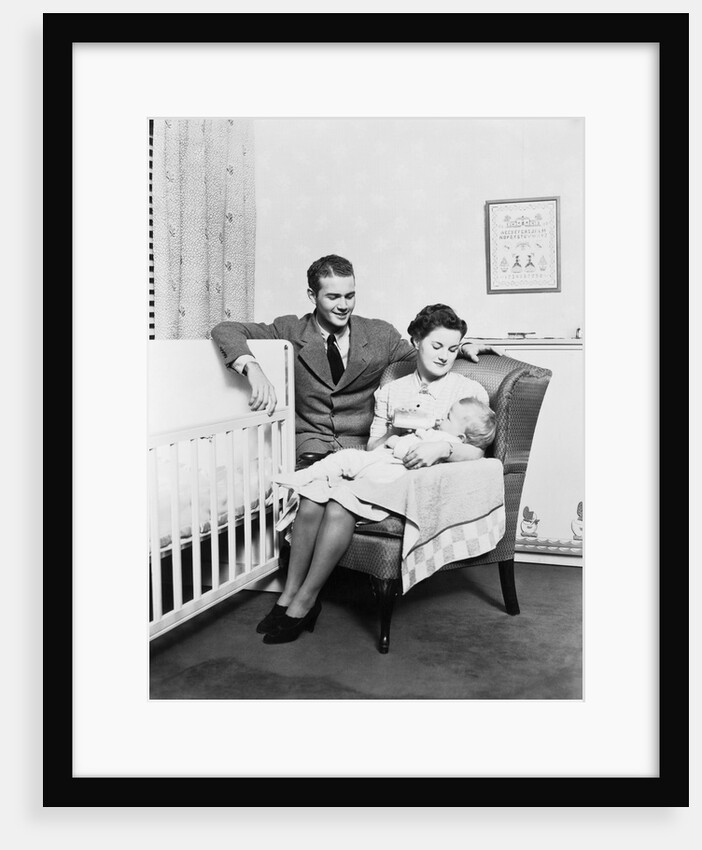 1930s 1940s mother sitting in chair in nursery baby 11 months on lap drinking from bottle father looking on leaning on crib by Anonymous