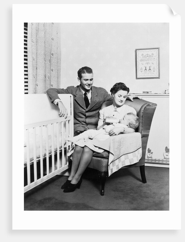 1930s 1940s mother sitting in chair in nursery baby 11 months on lap drinking from bottle father looking on leaning on crib by Anonymous