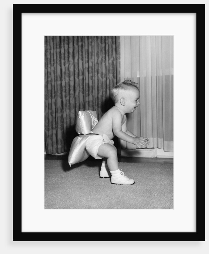 1950s laughing baby in diaper and shoes learning to walk with a pillow tied to his rear end by Anonymous