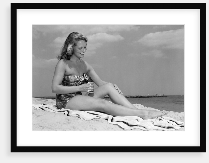 1950s woman in strapless one-piece bathing suit seated on beach towel putting on suntan lotion by Anonymous