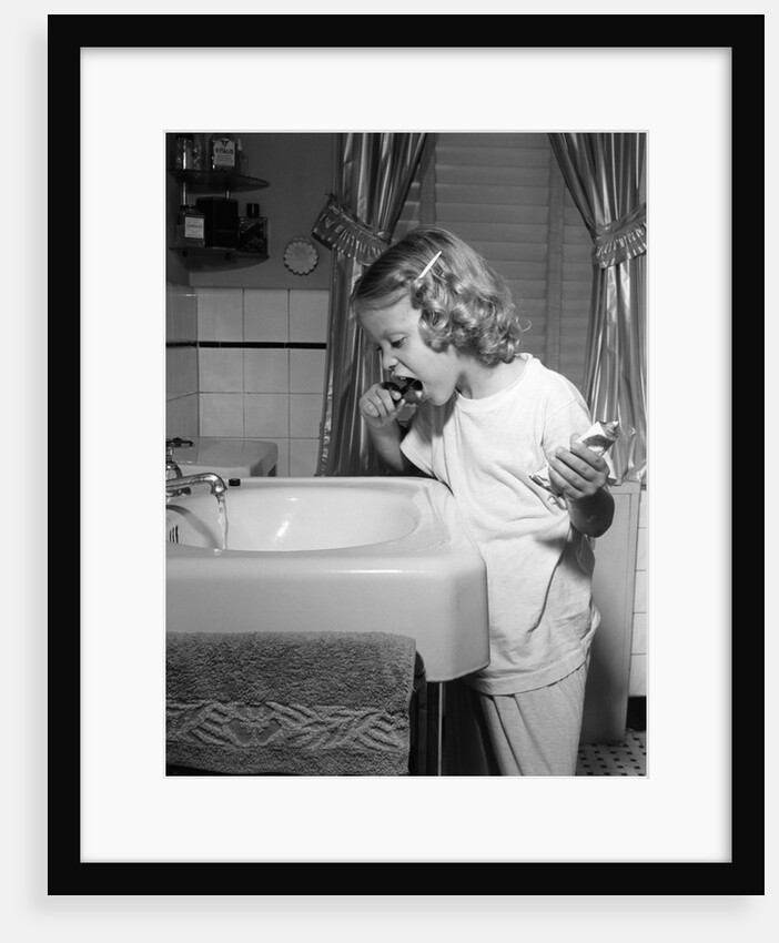 1950s little blond girl in pajamas standing at bathroom sink brushing her teeth toothpaste and toothbrush in hand by Anonymous