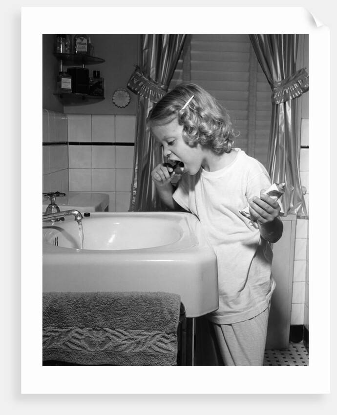 1950s little blond girl in pajamas standing at bathroom sink brushing her teeth toothpaste and toothbrush in hand by Anonymous