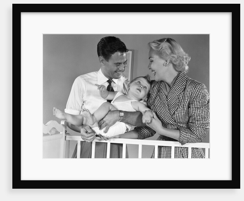 1950s smiling parents picking baby up out of crib by Anonymous