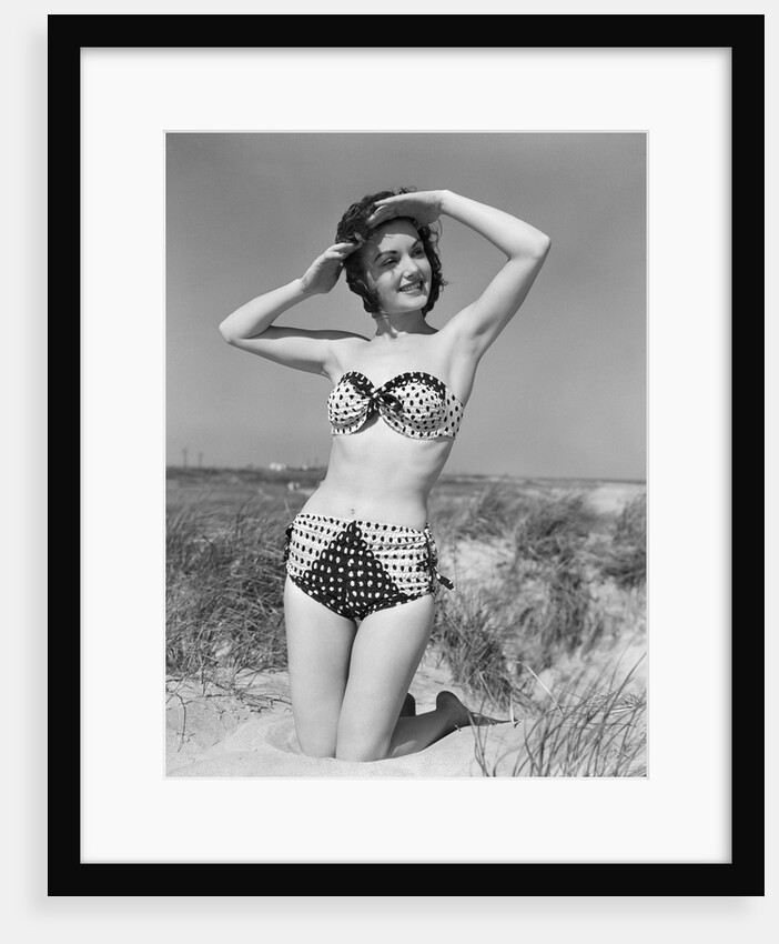 1950s smiling young woman kneeling in grassy sand wearing polka dot bikini shading eyes from sun by Anonymous