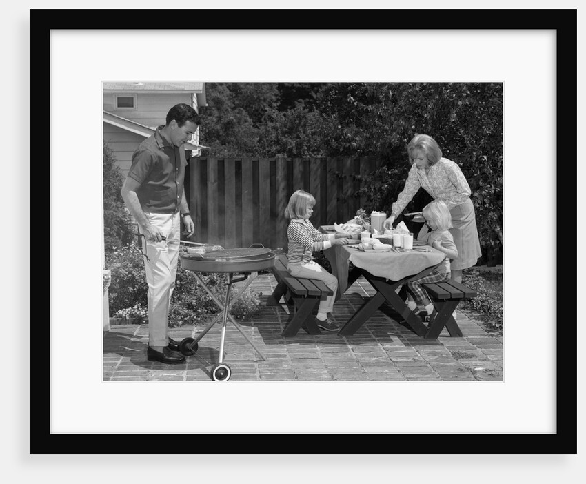 1960s man on patio grilling steak with 2 daughters seated at picnic table & wife standing serving food by Anonymous