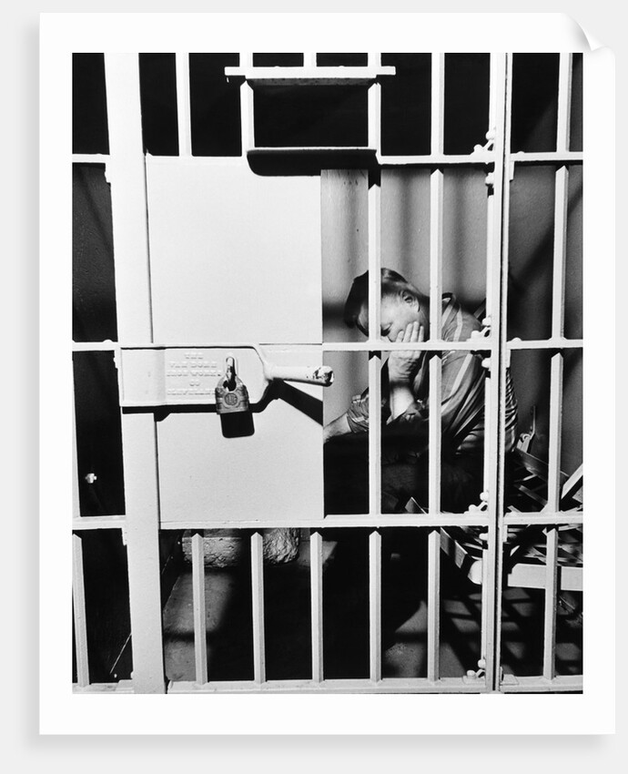 1960s man seated on bench in jail cell with head in hand looking downward by Anonymous