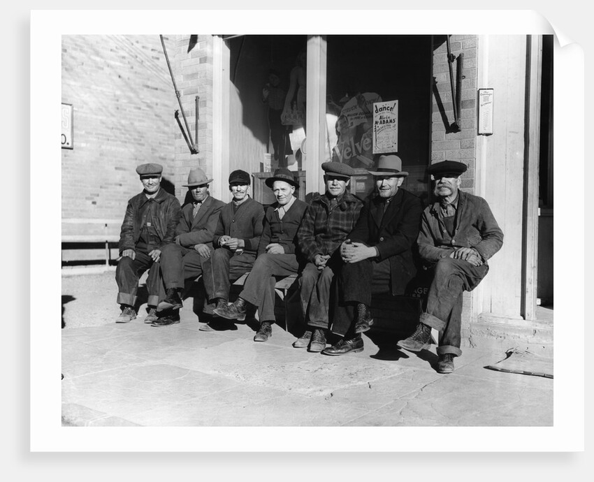 1920s 1930s group of old men in hats & dungarees sitting on bench in front of store window looking at camera by Anonymous
