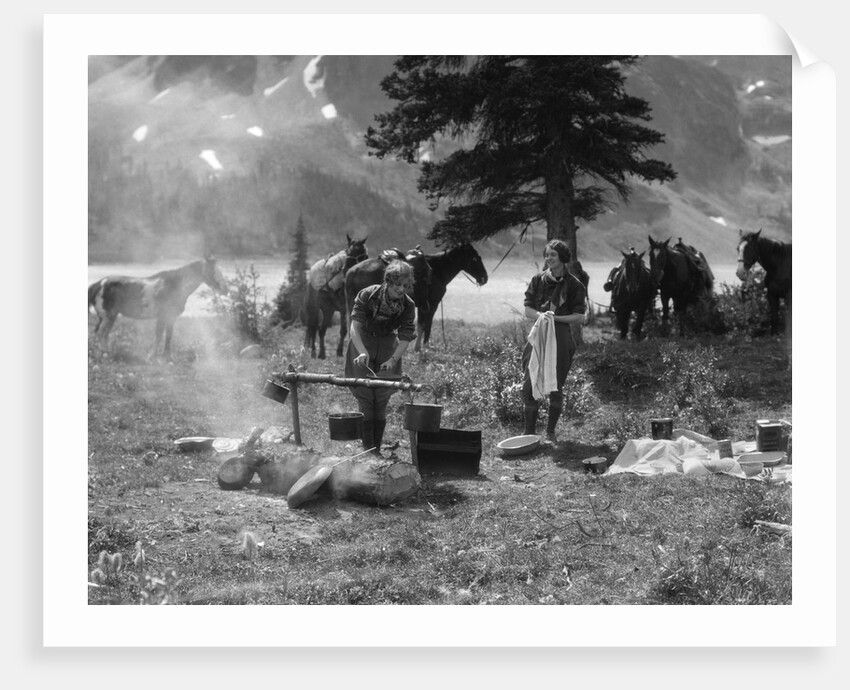 1920s 1930s two women at campsite woman cooking preparing food over campfire horses with riding gear in background by Anonymous