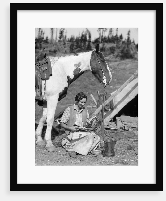 1920s 1930s woman sitting outside of tent next to pinto horse pouring syrup from tin onto pancakes wearing vest and chaps glacier park by Anonymous