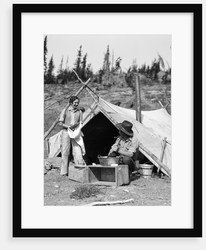 1930s smiling talking couple working by rustic western campsite tent man in cowboy hat smoking pipe washing skillet woman drying dishes by Anonymous