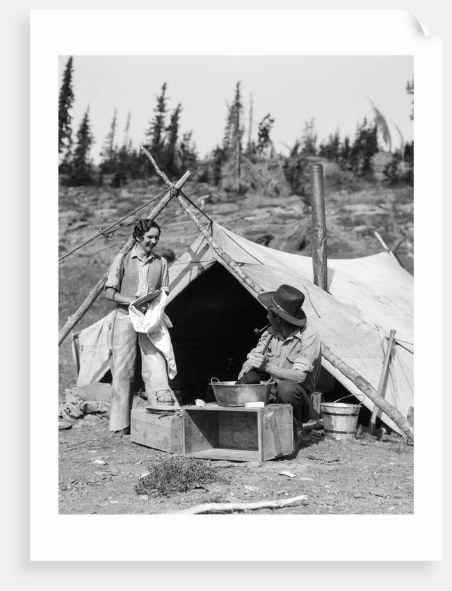 1930s smiling talking couple working by rustic western campsite tent man in cowboy hat smoking pipe washing skillet woman drying dishes by Anonymous