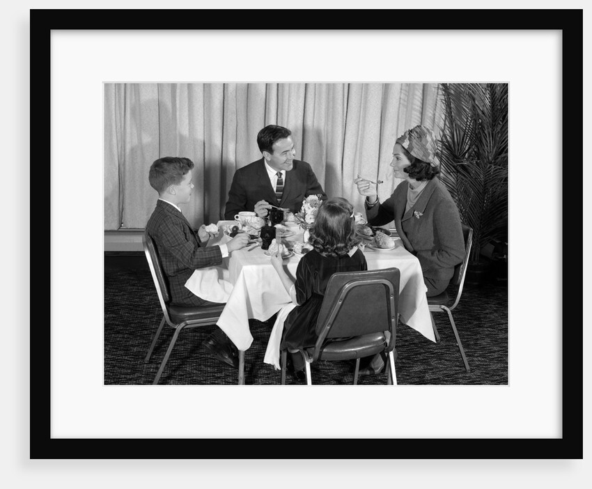 1960s smiling happy family of four eating meal in restaurant dad talking to mom who is wearing turban style hat by Anonymous