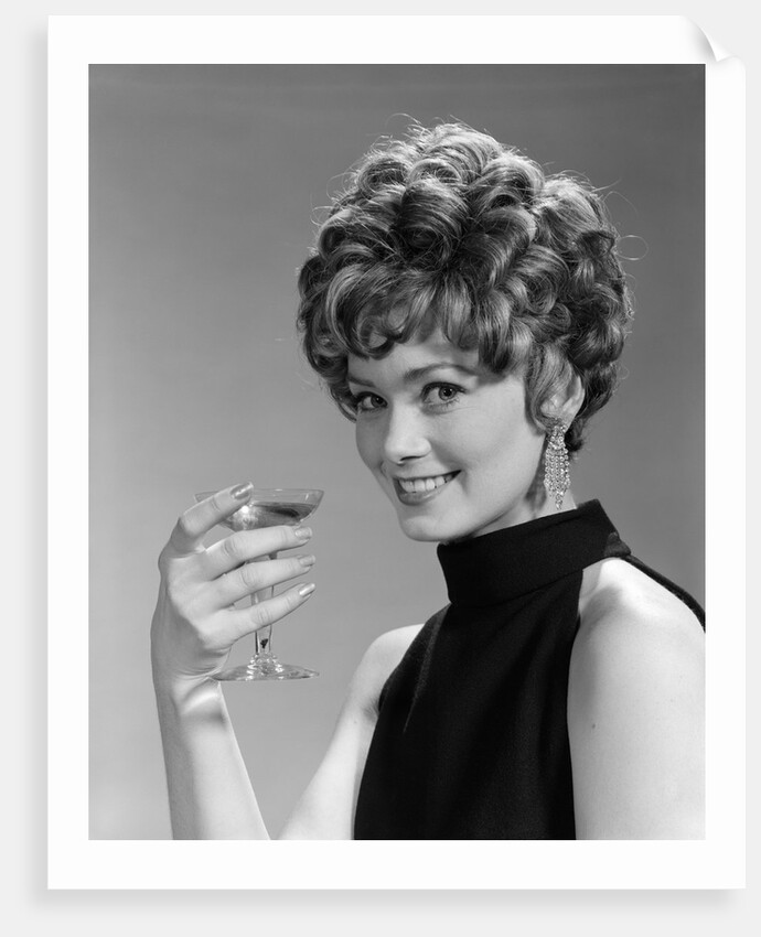 1960s glamorous woman with curly hair and dangling earrings smiling holding a champagne glass looking at camera by Anonymous