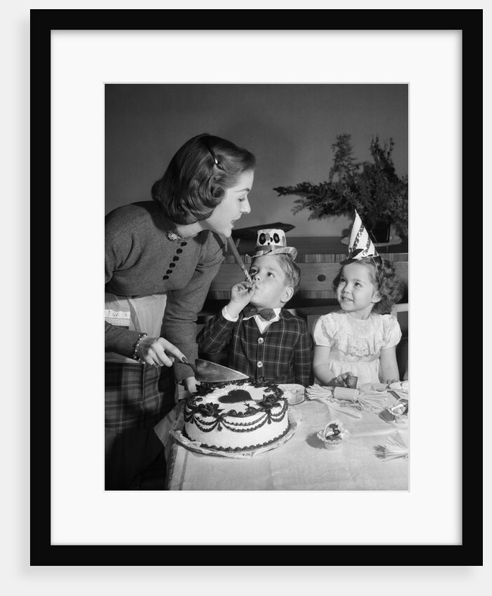 1950s woman mother cutting birthday cake for two children sitting at table wearing party hats by Anonymous