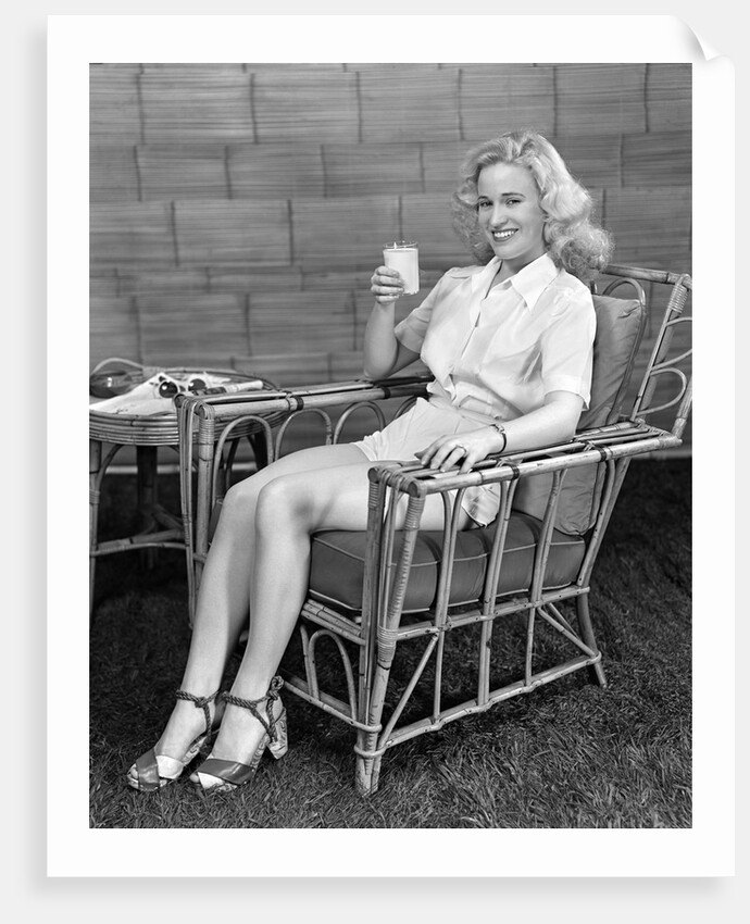 1940s blonde woman in bamboo chair holding up glass of milk smiling wearing white shorts looking at camera by Anonymous